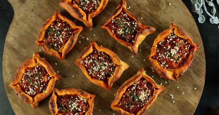 Small open-faced savoury pastries topped with spiced minced meat and sesame seeds, arranged on a round wooden board.