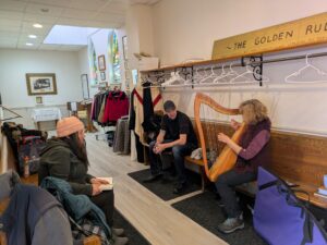 Iris Jones, a volunteer at St. Thomas parish, plays the harp for two Open Hearts Café clients, Jason and Joyce, in a hallway with supplies and clothing for those in need. Photo: Ann Marie Taylor